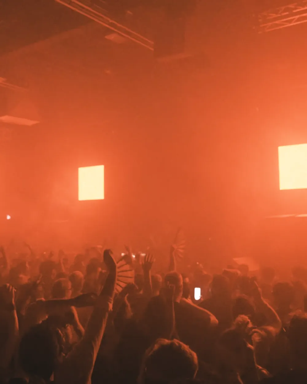 Hands in the air across a crowded dance floor in orange haze at Demo Room